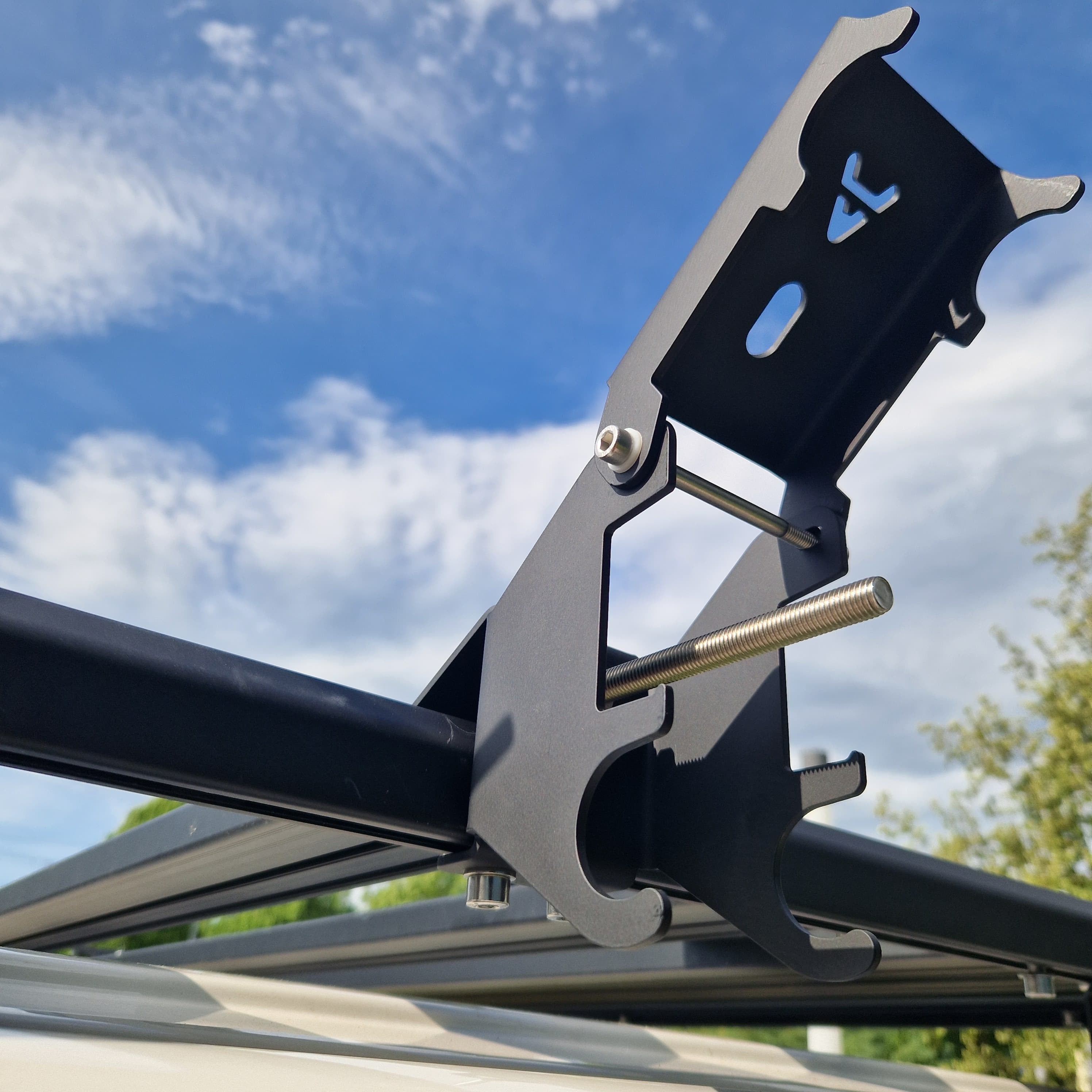 Aufgenommen im Freien: Der Airholder Wagenheberhalter ist hier in geöffneter Position zu sehen – die Halteplatte steht hoch, die Spannspindel ist ausgefahren und der Zugang zum Einlegen/Entnehmen des Hi-Lift ist frei. Die pulverbeschichtete Edelstahlkonstruktion sitzt fest am Außenprofil des Airholder Dachträgers und ist für schnelles Handling beim Offroad-Einsatz ausgelegt. Position: seitlich am Außenrahmen des Dachträgers (links/rechts montierbar).