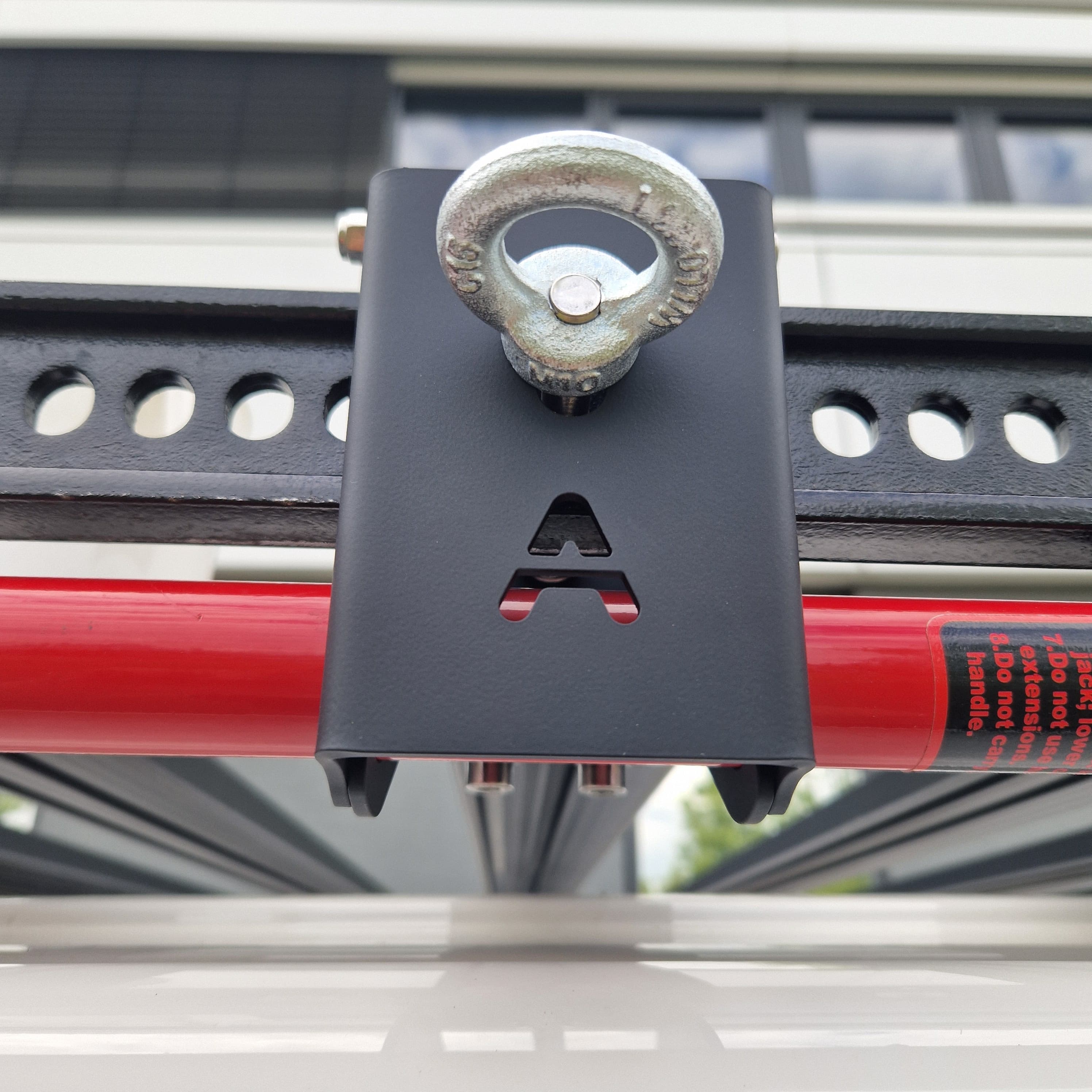 Outdoor-Aufnahme im Detail: Der Airholder Wagenheberhalter aus Edelstahl mit matter, widerstandsfähiger Pulverbeschichtung sitzt am seitlichen Außenprofil des Dachträgers. Die Halterplatte mit Airholder-Ausschnitt fixiert den roten Hi-Lift-Wagenheber spielfrei, während die integrierte Ringöse oben als zusätzlicher Sicherungspunkt für Gurt, Karabiner oder Vorhängeschloss dient. Position: seitlich am Außenrahmen des Airholder Dachträgers (links/rechts montierbar).