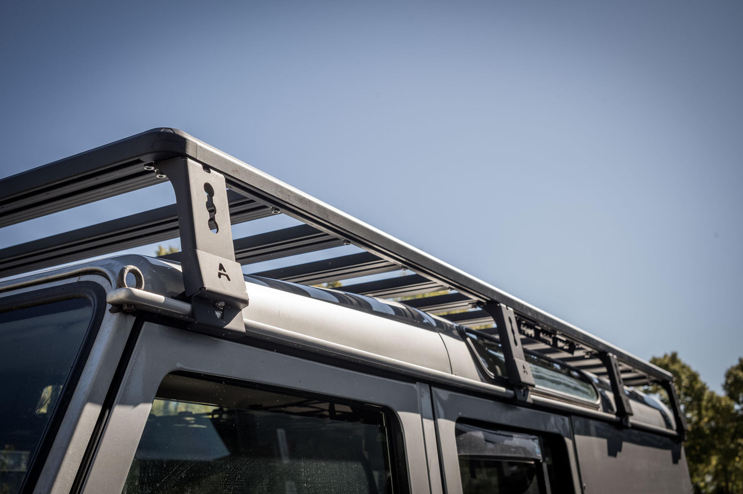 Außenaufnahme bei klarem Himmel, aufgenommen aus schräger Untersicht entlang der Dachkante: Zu sehen ist die Dachplattform (Roof Rack) auf einem Land Rover Defender 110 mit den Querstreben und dem umlaufenden Rahmen. Im Fokus stehen die seitlichen Halterungen mit Schlüsselloch-Ausführung und „A“-Markierung, die die Plattform am Fahrzeugdach abstützen. Die Perspektive zeigt besonders gut die stabile Rahmenkonstruktion und die offene Bauweise für flexible © Thomas A. Berger | www.diebergerdesigner.de