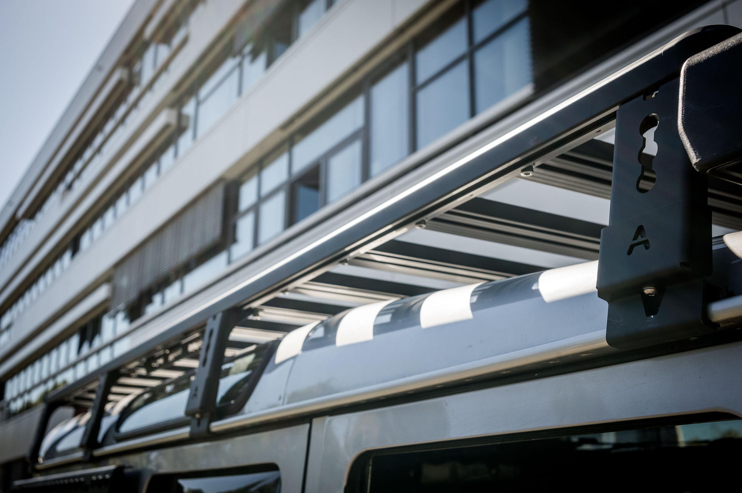 Außenaufnahme in starker Untersicht entlang der Dachkante: Der Fokus liegt auf der Dachplattform (Roof Rack) eines Land Rover Defender 110 mit den Querstreben und den seitlichen Halterungen, die am Fahrzeugdach befestigt sind. Im Vordergrund ist ein Befestigungsbügel mit „A“-Markierung zu sehen, während die Lamellen/Querträger der Plattform nach hinten verlaufen. Im Hintergrund erkennt man die Glasfassade eines modernen Gebäudes, wodurch die Szene klar © Thomas A. Berger | www.diebergerdesigner.de