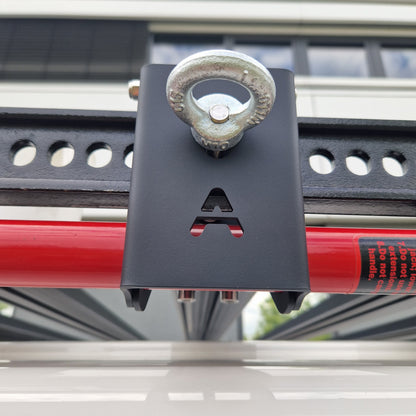 Outdoor-Aufnahme im Detail: Der Airholder Wagenheberhalter aus Edelstahl mit matter, widerstandsfähiger Pulverbeschichtung sitzt am seitlichen Außenprofil des Dachträgers. Die Halterplatte mit Airholder-Ausschnitt fixiert den roten Hi-Lift-Wagenheber spielfrei, während die integrierte Ringöse oben als zusätzlicher Sicherungspunkt für Gurt, Karabiner oder Vorhängeschloss dient. Position: seitlich am Außenrahmen des Airholder Dachträgers (links/rechts montierbar).
