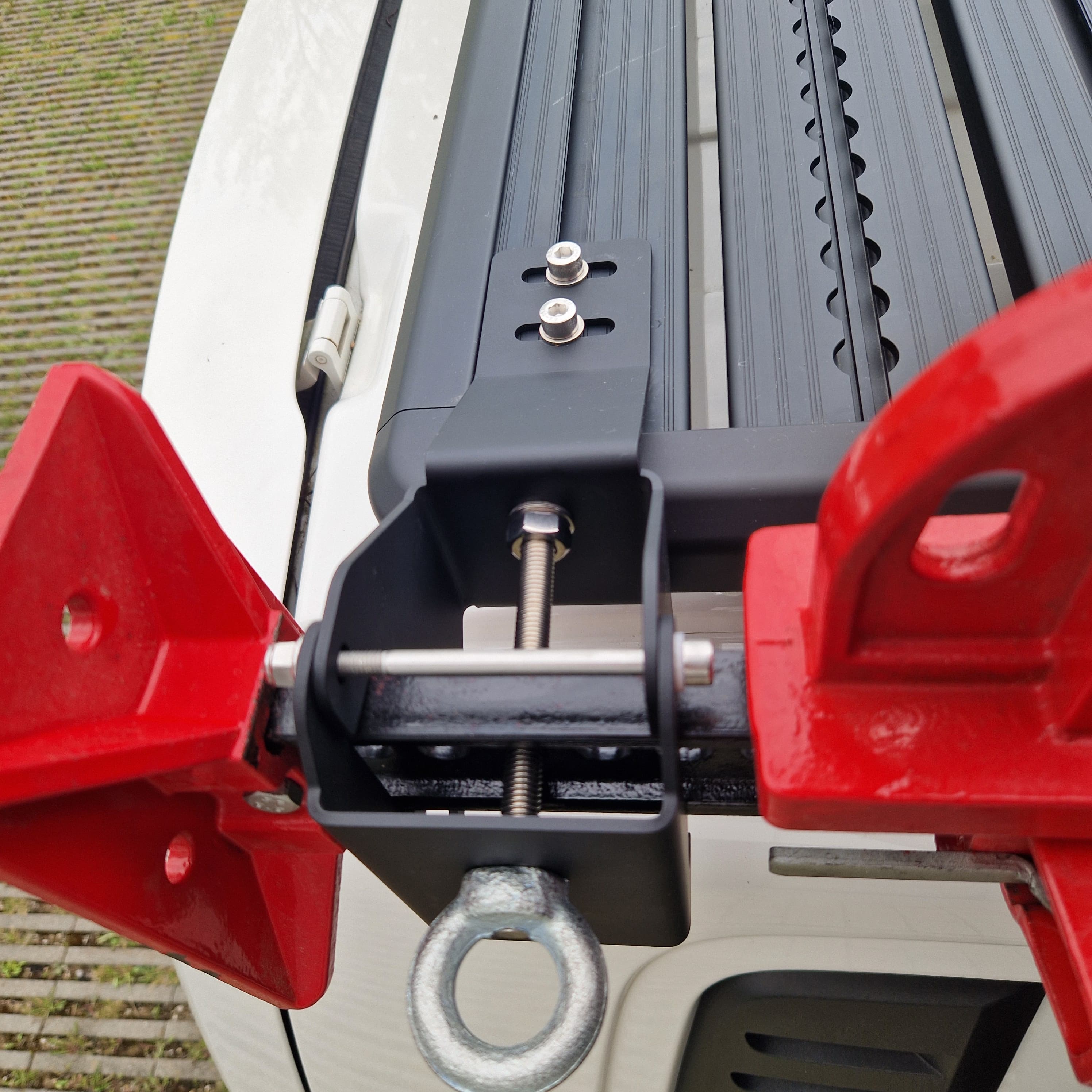 Aufnahme im Freien von oben: Der Airholder Wagenheberhalter sitzt am seitlichen Außenprofil und klemmt den Hi-Lift über eine massive Schraubklemmung sicher ein. Die Edelstahl-Hardware und die pulverbeschichtete Halterung sorgen für festen Sitz, auch bei ruckeligen Strecken. Unten dient die Ringöse als zusätzlicher Sicherungspunkt für Spanngurt oder Schloss. Position: Seitenkante des Dachträgers am Außenrahmen (links/rechts montierbar).
