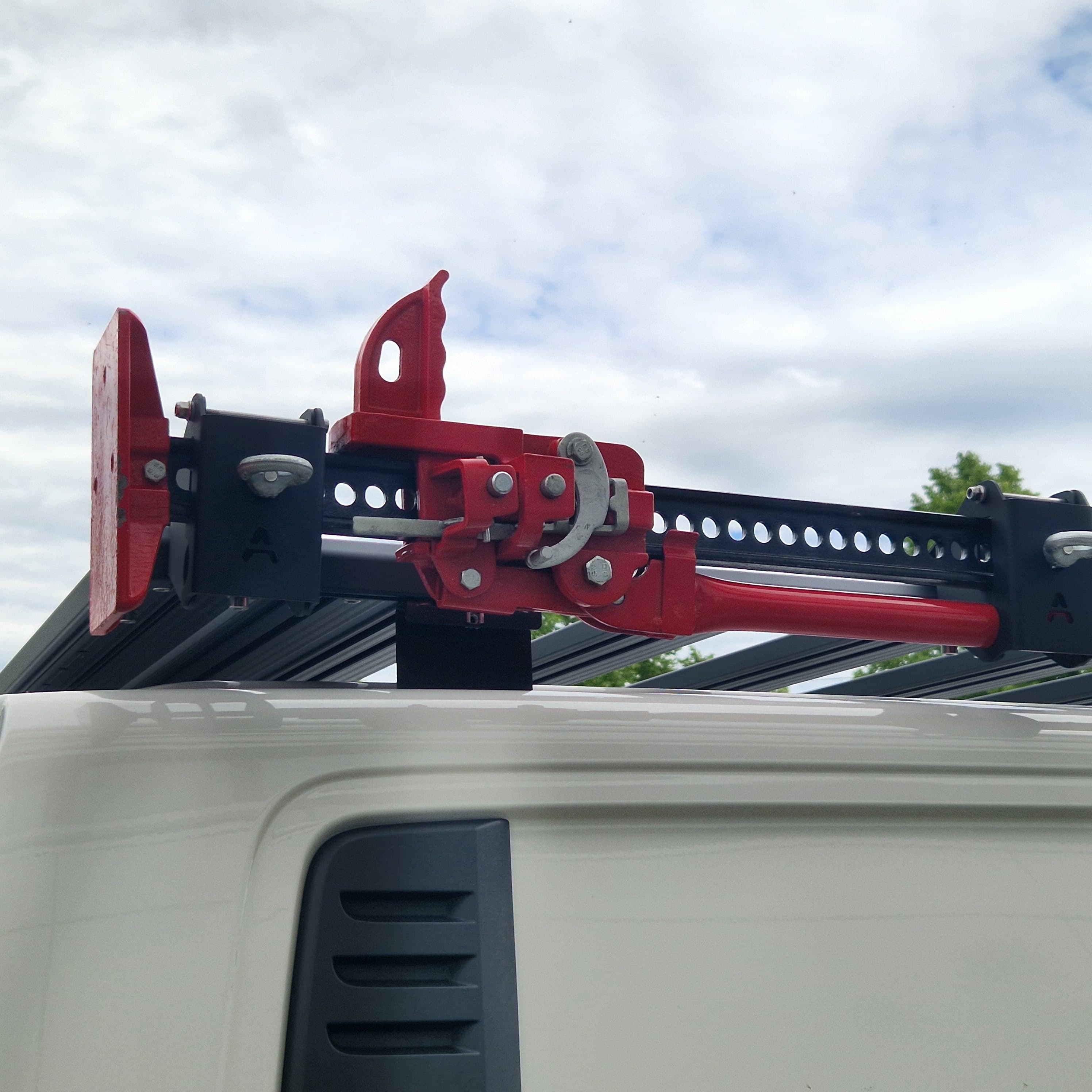 Aufgenommen im Freien: Ein rot pulverbeschichteter Wagenheber (Hi-Lift-Typ) ist seitlich am Airholder Dachträger montiert. Der Halter aus rostfreiem Stahl sitzt am Außenprofil und fixiert den Wagenheber über eine stabile Klemmmechanik und eine Lochleiste/Schiene sicher gegen Klappern und Vibrationen. So bleibt der Wagenheber schnell zugänglich, ohne die Ladefläche oben zu blockieren. Position: Seitenkante des Dachträgers am Außenprofil (links oder rechts montierbar).
