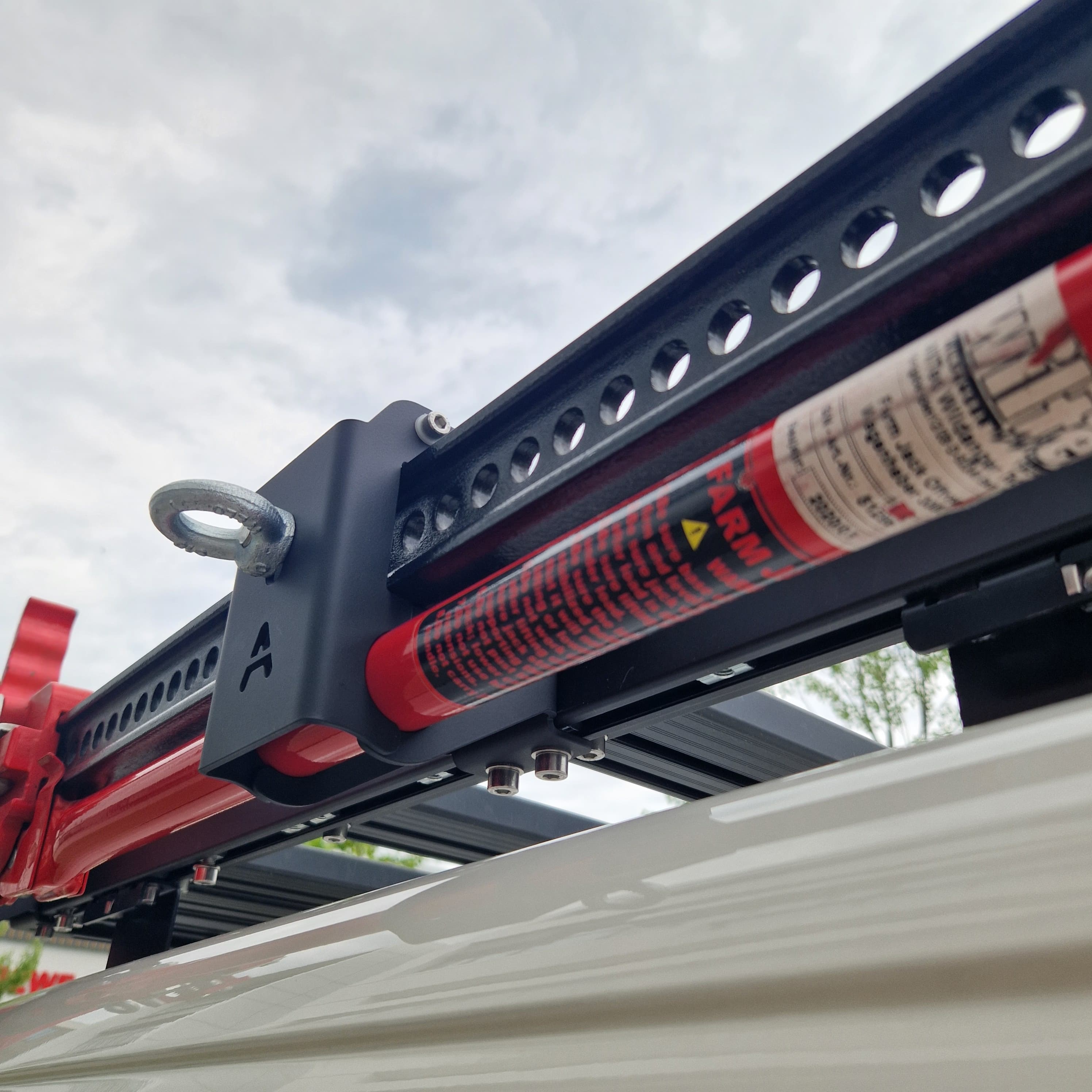 Outdoor-Perspektive von unten: Der rote Hi-Lift-Wagenheber läuft parallel zum Airholder Außenprofil und wird von einem pulverbeschichteten Edelstahlhalter sauber geführt und festgeklemmt. Die Lochleiste/Schiene am Wagenheber sowie die seitliche Ringöse am Halter bieten zusätzliche Möglichkeiten zum Sichern und Verzurren. Position: seitliche Unterkante am Außenrahmen des Airholder Dachträgers (links/rechts montierbar).
