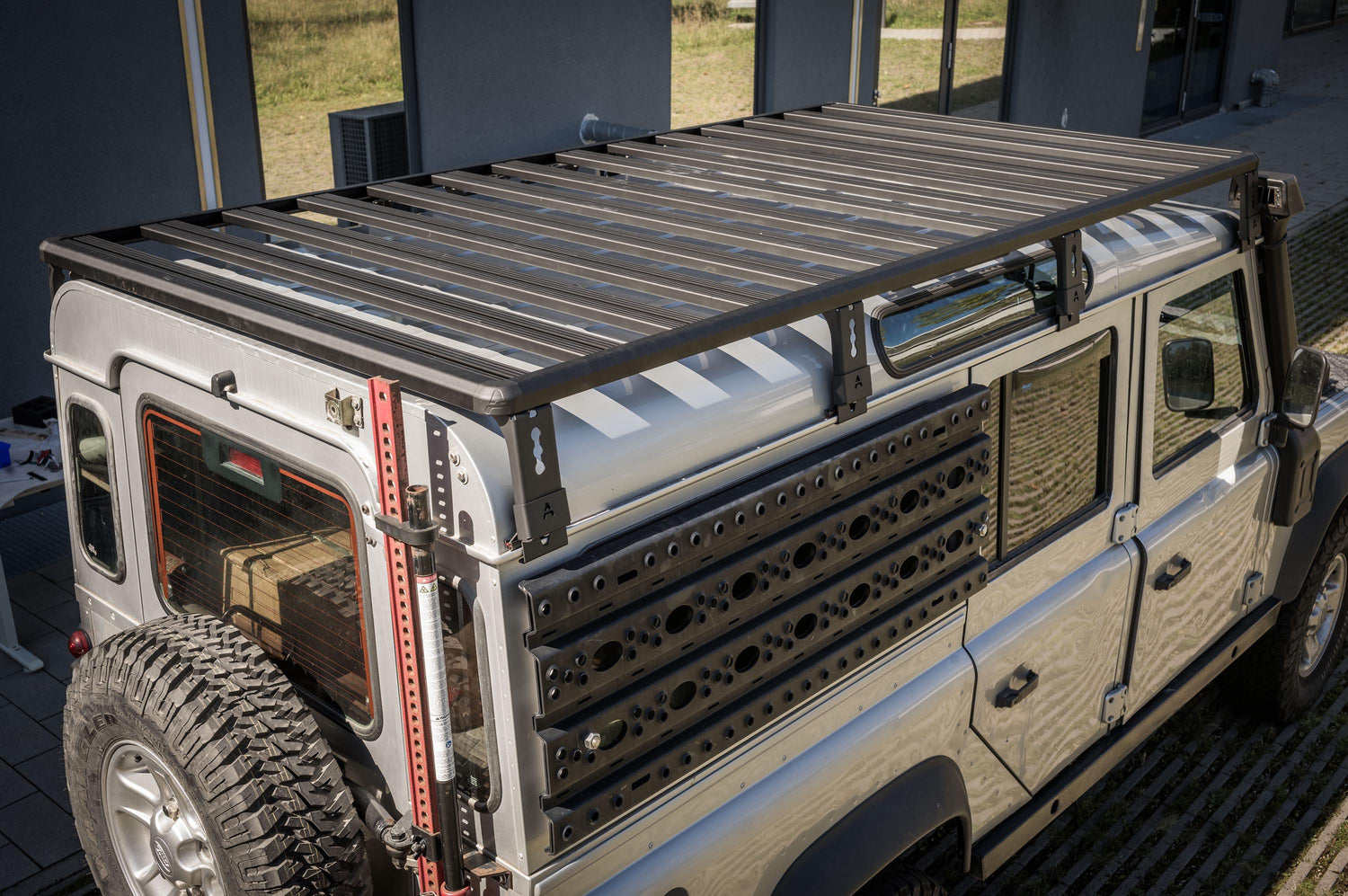 Schrägaufnahme von oben hinten links: Ein Land Rover Defender 110 steht vor einem Gebäude und ist mit einer großflächigen Dachplattform (Roof Rack) mit Querstreben ausgestattet. An der Fahrzeugseite sind robuste Anbauplatten/Recovery-Boards montiert, am Heck sind Reserverad und ein Hi-Lift-/Wagenheber befestigt – ein typisches Setup für Offroad und Overlanding.

© Thomas A. Berger | www.diebergerdesigner.de 