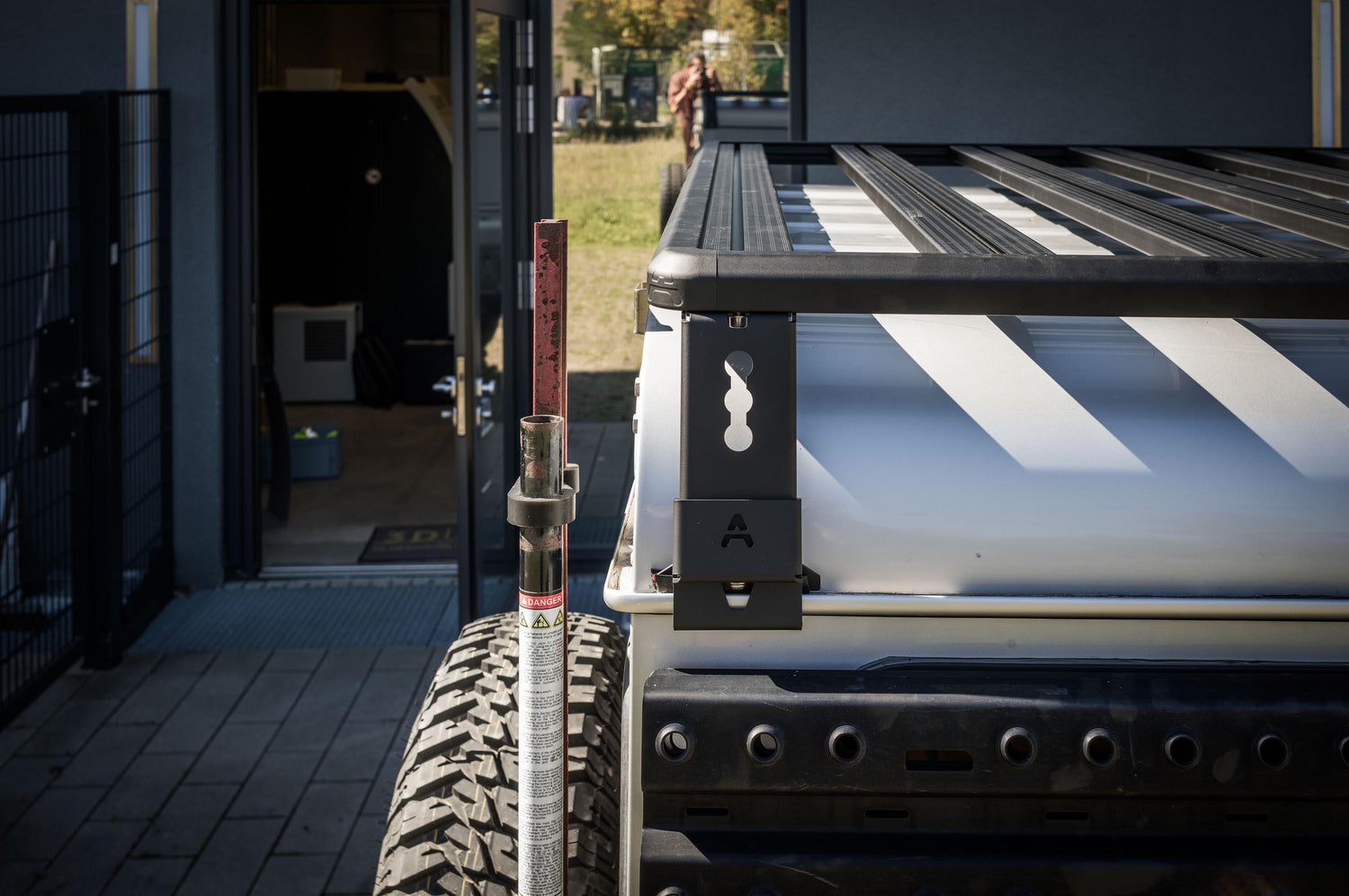 Außenaufnahme im Heckbereich eines Land Rover Defender 110: Im Vordergrund ist die Ecke der Dachplattform (Roof Rack) mit den Querstreben zu sehen, inklusive der seitlichen Halterung mit „A“-Markierung und Schlüsselloch-Ausführung. Daneben steht ein Hi-Lift-/Wagenheber am Fahrzeug, während darunter die gelochte Seitenplatte für Offroad-Zubehör sichtbar ist. Die Szene ist bei Tageslicht vor einem Gebäude aufgenommen und zeigt die stabile, kantige Rahmenkonstrukti © Thomas A. Berger | www.diebergerdesigner.de