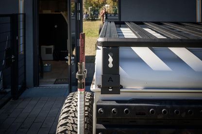 Außenaufnahme im Heckbereich eines Land Rover Defender 110: Im Vordergrund ist die Ecke der Dachplattform (Roof Rack) mit den Querstreben zu sehen, inklusive der seitlichen Halterung mit „A“-Markierung und Schlüsselloch-Ausführung. Daneben steht ein Hi-Lift-/Wagenheber am Fahrzeug, während darunter die gelochte Seitenplatte für Offroad-Zubehör sichtbar ist. Die Szene ist bei Tageslicht vor einem Gebäude aufgenommen und zeigt die stabile, kantige Rahmenkonstrukti © Thomas A. Berger | www.diebergerdesigner.de