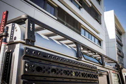 Außenaufnahme in niedriger Perspektive: Zu sehen ist die hintere Seitenpartie eines Land Rover Defender 110 mit montierter Dachplattform (Roof Rack). Der Fokus liegt auf den Halterungen und der Rahmenkonstruktion des Dachträgers sowie den seitlich befestigten, gelochten Anbauplatten für Offroad-Equipment. Im Hintergrund ist die Fassade eines modernen Gebäudes zu erkennen, was die Aufnahme klar als Outdoor-Szene bei Tageslicht  Roof Rack Land Rover defender 110 © Thomas A. Berger | www.diebergerdesigner.de