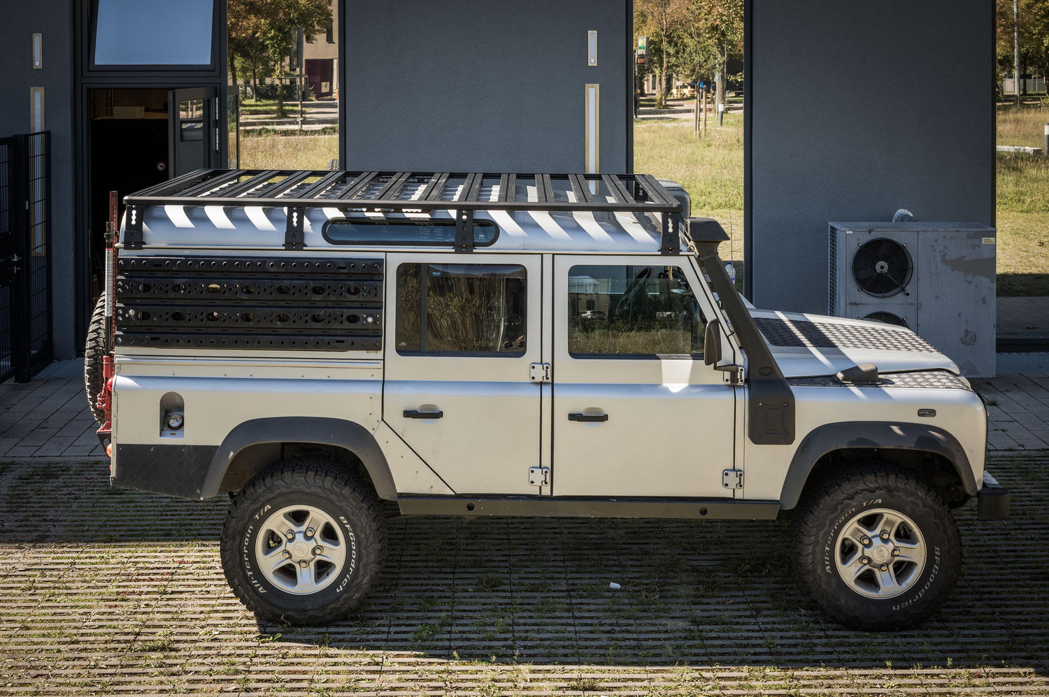Außenaufnahme bei Tageslicht: Ein Land Rover Defender 110 im Offroad-Setup steht im Freien vor einem Gebäude. Auf dem Dach ist eine großflächige Dachplattform (Roof Rack) mit Querstreben montiert, die über mehrere Halterungen am Fahrzeug befestigt ist. An der Fahrzeugseite sind robuste Anbauplatten/Recovery-Boards angebracht, außerdem ist ein Schnorchel am vorderen Kotflügel sichtbar. So wird die durchgehende Ladefläche des DachRoof Rack Land Rover defender 110
© Thomas A. Berger | www.diebergerdesigner.de 